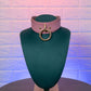 Pink leather choker with gold ring on a green mannequin against a textured brick wall.