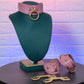 Pink leather choker and cuffs on a green mannequin bust with a gold key against a textured purple and blue background.