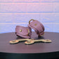 Pink leather handcuffs with gold hardware on a dark surface against a textured light blue background
