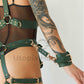Wide chest harness with bondage garters with handcuffs attached to the garters with D-rings. The handcuffs are fastened to the D-Rings on the garters. Close-up view. Green leather. Gold hardware.