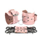 Pink leather handcuffs with two connectors and carabiners lie side by side, one handcuff is shown from the side of the buckle and lies half sideways the second handcuff stands half sideways and is shown from the side of the D-ring for which the carabiner is fixed. Dark nickel hardware. 