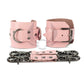Pink leather handcuffs with two connectors with carabiners are lying side by side, one handcuff is shown with a buckle and it stands on a rib, the second handcuff is shown with a D-ring for which the carabiners are attached and it also stands on a rib. Front view. Dark nickel hardware. 